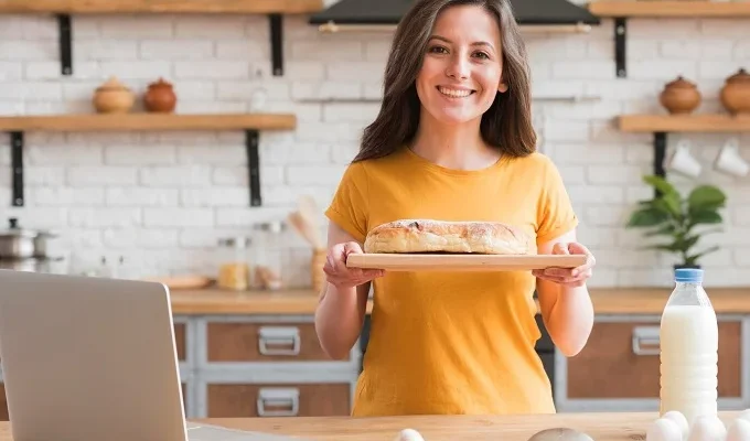 Smiling woman in a kitchen holding freshly baked bread, promoting her home-based bakery business.