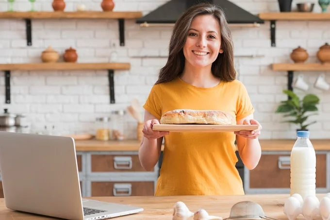 Smiling woman in a kitchen holding freshly baked bread, promoting her home-based bakery business.